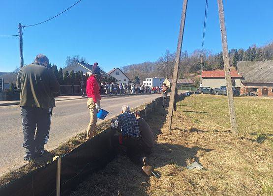 Po prawej stronie drogi kilka osób wykonuje montaż ogrodzenia naprowadzającego dla plazów. Po lewej